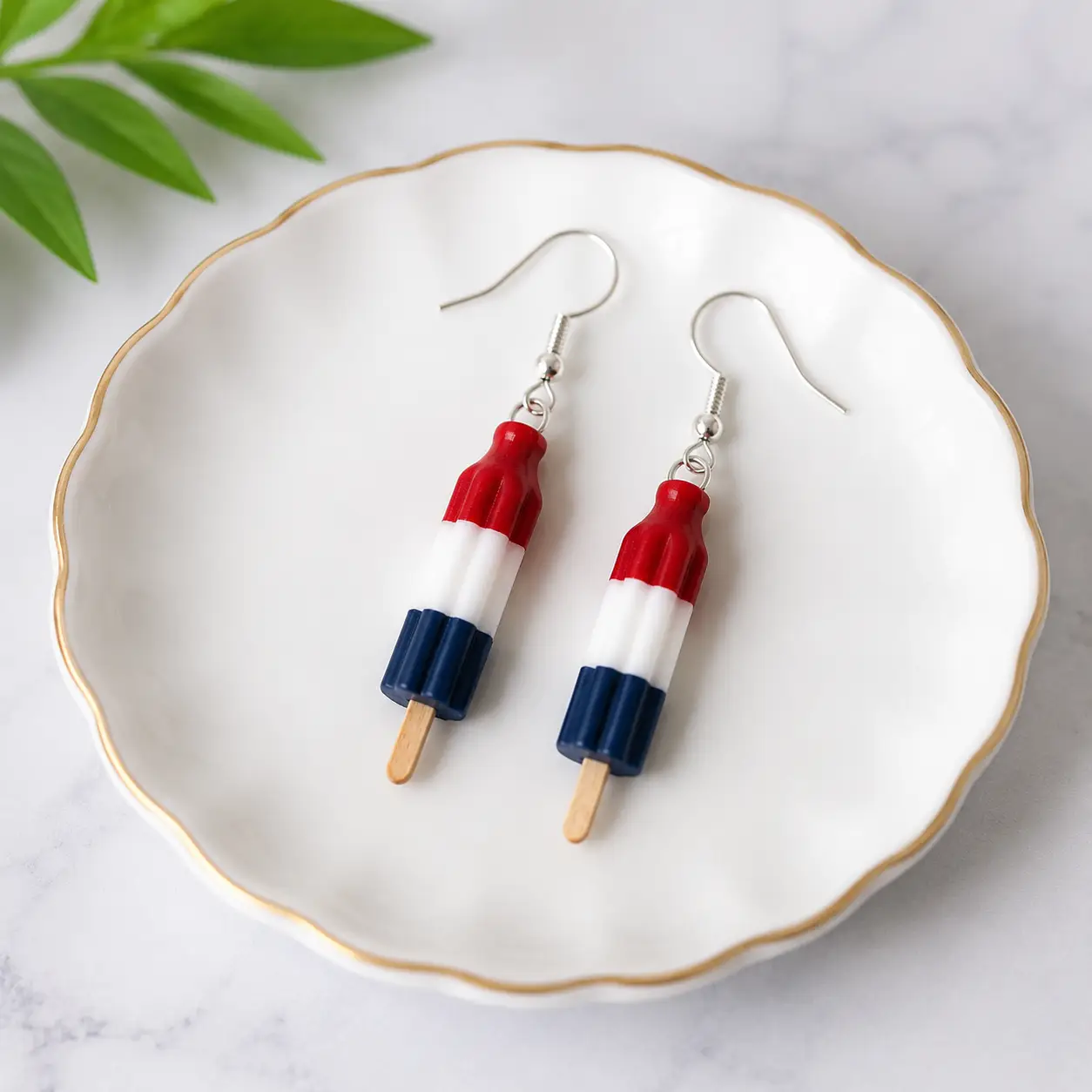 Red, white, and blue popsicle earrings resting on a white ceramic plate with gold trim.