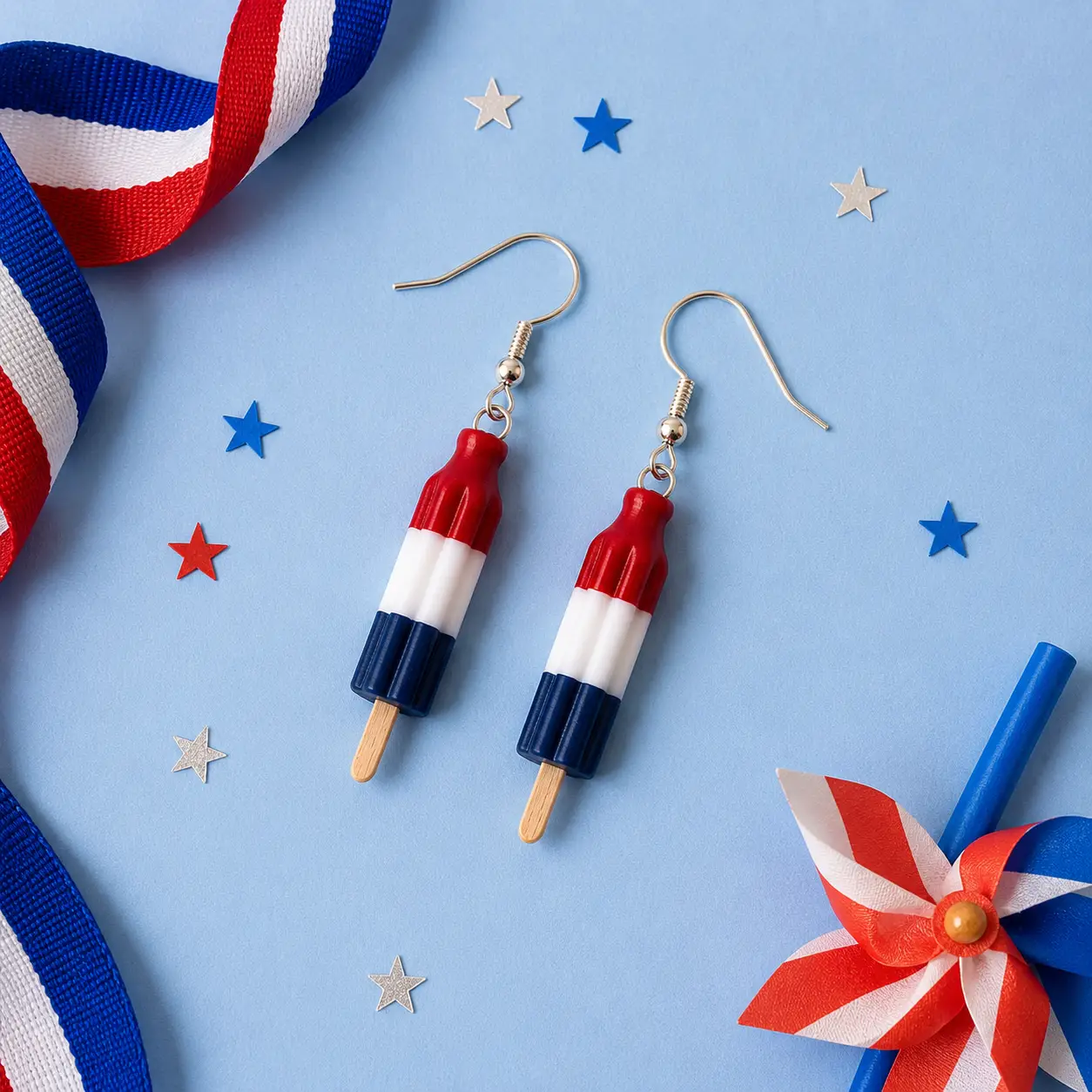 Bomb Pop earrings displayed on a blue background with patriotic ribbon, stars, and pinwheel decorations.